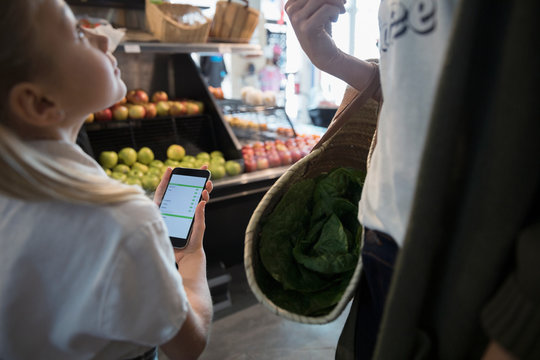 Mother And Daughter With Smart Phone List Grocery Shopping In Produce Section At Market