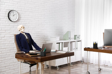 Human skeleton in suit using laptop at table in office