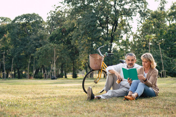Caucasian elderly couple relaxing and sitting at the park reading the book while husband looking to...
