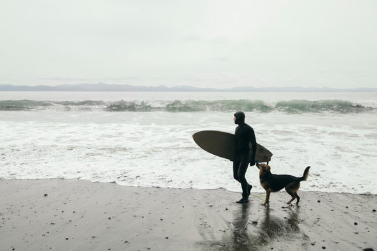 Male surfer with dog carrying surfboard on rugged beach