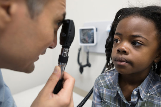 Male doctor using otoscope, examining eye of girl patient in clinic exam room