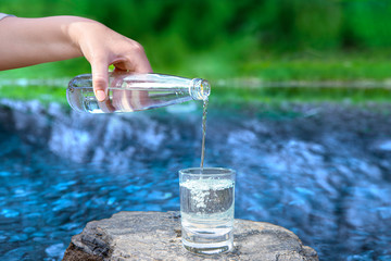 Female hand pours pure spring water from bottle into glass stan on background of mountain river. Hand holding drinking water glass.