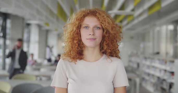 Front view of curly red haired manager standing in modern office workspace. Portrait of attractive smiling young woman business coach looking to camera