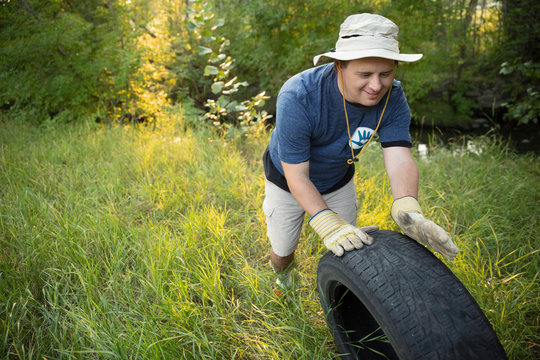 Man with down syndrome volunteering, cleaning up garbage in woods, rolling tire in grass