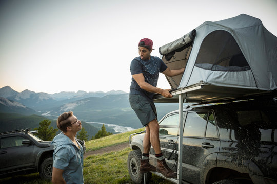 Male friends camping, climbing up into SUV rooftop tent in mountain field, Alberta, Canada