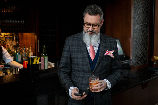 Stylish hipster businessman in suit drinking cocktail and checking smart phone in bar