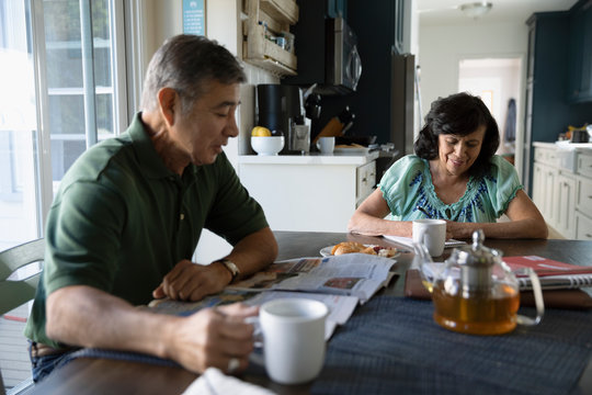 Latinx senior couple reading newspaper and enjoying breakfast at kitchen table