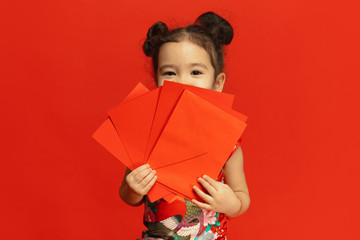 Holding red envelopes, looks happy, smiling. Happy Chinese New Year 2020. Asian cute little girl isolated on red background in traditional clothing. Celebration, human emotions, holidays. Copyspace.