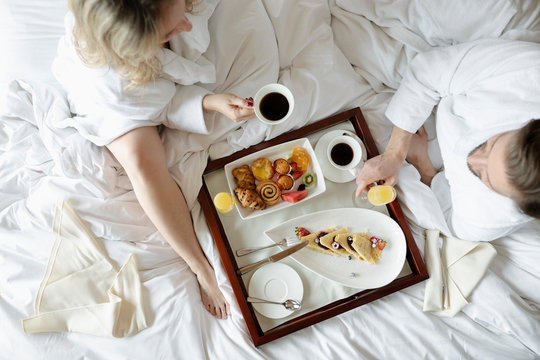 Couple in bathrobes enjoying breakfast in bed in luxury hotel, enjoying romantic weekend