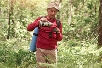 Outdoor shot of old man with bag on back, holding thermos and being ready to drik hot tea or...