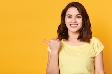 Horizontal shot of attractive young female pointing aside with cheerful expression, showing something amazing, posing isolated over yellow backgroud. Copy space foer advertisment or promotional text.
