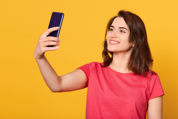 Horizontal shot of pretty girl taking self portrait with her smart phone, adorable lady looking devie's at camera with charming smile, attractive model posing isolated over yellow studio background.