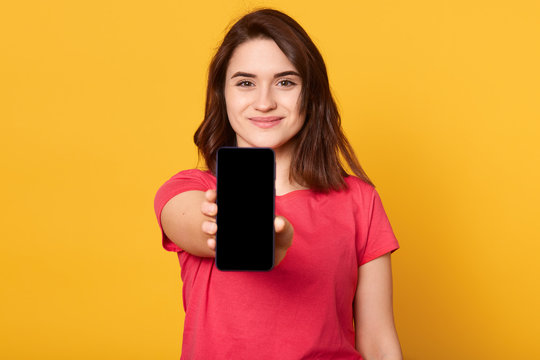 Close up portrait of young woman showing display of mobile cell phone with black blank screen and looks smiling directly at camera isolated over yellow background. Copy space for advertisment.
