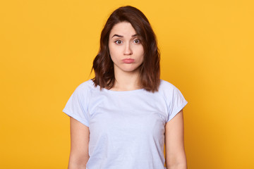 Obraz premium Studi oshot of dark haired young woman dresses casual white t shirt, standing and looking at camera, posing with pouty lips isolated over yellow background, being offended and having bad mood.