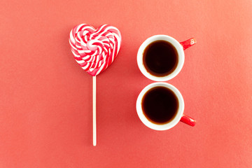 Two red cups of hot espresso, coffee or tea and a lollipop in the shape of a heart on bright pink background. Creative idea for romantic date for Valentines Day with coffee and sweets, flat lay