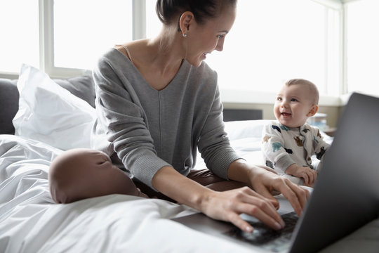 Mother With Baby Son Working From Home, Using Laptop On Bed