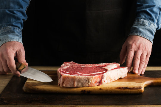 Hombre preparando chulet&oacute;n de vaca con cuchilla sobre tabla de madera r&uacute;stica. Vista de frente