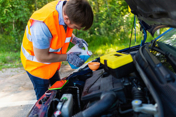 Mechanic in an orange vest pours liquid into the tank in the engine. A man in gloves fills the coolant in the tank radiator cap. Planned maintenance of the vehicle