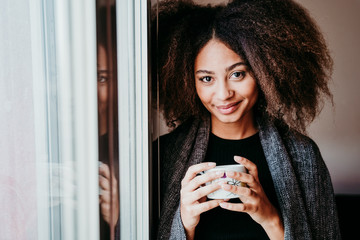 portrait of beautiful afro american young woman by the window holding a cup of coffee. Lifestyle indoors