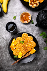 Healthy breakfast with fresh hot waffles hearts, pancakes flowers with berry honey and exotic fruits on gray background, top view, flat lay. Food concept.