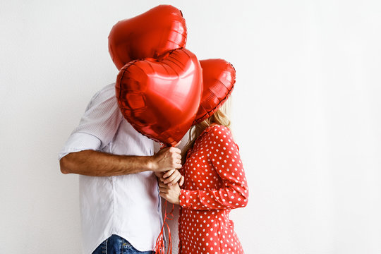 Couple. Love. Valentine's day. Emotions. Man is giving heart-shaped balloons to his woman, both smiling; on a white background