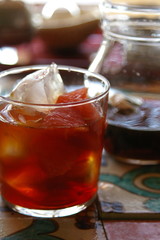 Iced tea in a glass with fruit and ice cube, with a jug of tea behind the glass.