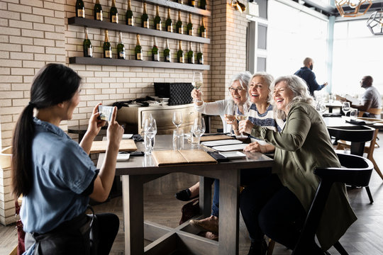 Waitress photographing happy women friends drinking white wine in wine bar
