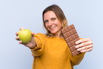 Blonde woman over isolated blue background taking a chocolate tablet in one hand and an apple in...