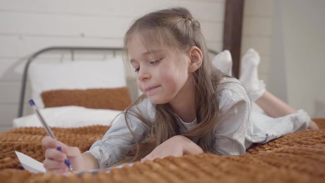 Portrait of charming Caucasian girl lying on bed and writing. Intelligent child showing i got an idea gesture and noting her thoughts. Lifestyle, relaxation, hobby.