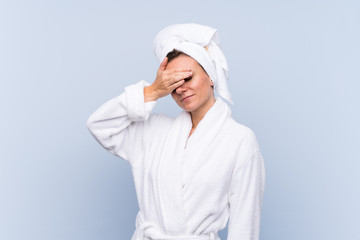 Woman in bathrobe over isolated blue background covering eyes by hands