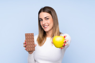 Young blonde woman over isolated blue background taking a chocolate tablet in one hand and an apple...