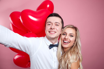 Beautiful young couple taking selfie on mobile phone and standing with red balloons on pink background