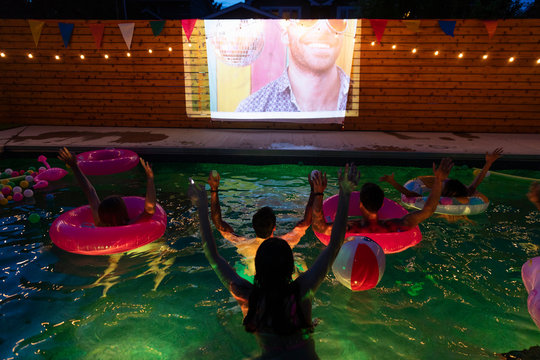 Happy Young Adult Friends In Summer Swimming Pool Cheering, Watching Movie On Projection Screen At Night