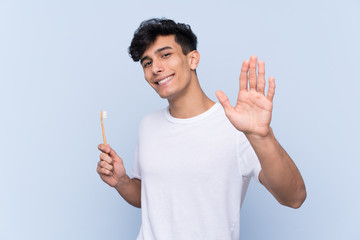 Young Argentinian man brushing his teeth over isolated blue background saluting with hand with happy expression