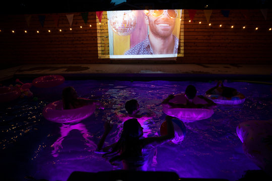 Young adult friends in summer swimming pool watching movie on projection screen at night