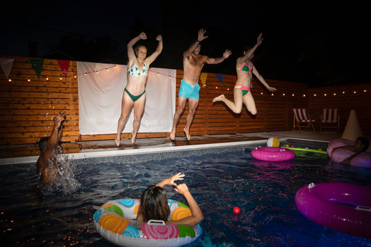 Playful young adult friends jumping into summer swimming pool at night