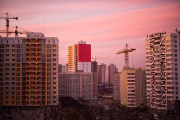 Industrial construction cranes and building silhouettes over sun at sunrise. Unfinished buildings development in pink sunset light. Silhouette of crane in morning light