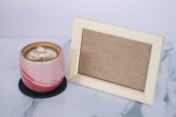 A pink ceramic cup of hot cappuccino and plain wooden frame on a marble table. Place text on wooden frame.