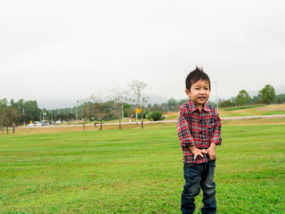 Asian cute child boy in red plaid and jeans standing and smiling outdoor with nature green glass background.