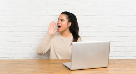 Young teenager Asian girl with a laptop in a table shouting with mouth wide open