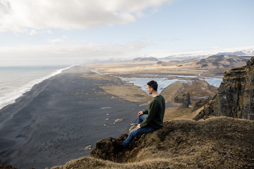 young male traveler sitting near a cliff view of a black beach and the Atlantic Ocean in Iceland