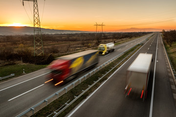Delivery trucks and cars in high speed driving on a highway through rural landscape. Fast blurred motion drive on the freeway. Freight scene on the motorway