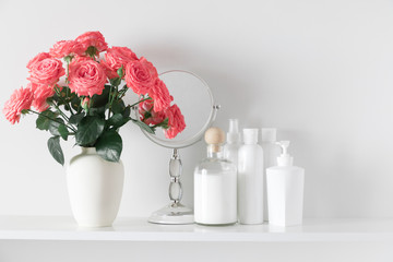 Soft light bathroom decor. Roses flowers of coral color in vase and mirror on shelf against white wall. Set of cosmetic bottles. Space for text.