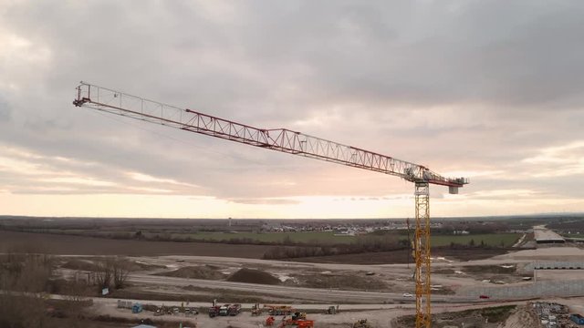 Drone footage of a construction crane on a construction site with a beautiful sunset with clouds. The camera is slowly moving above the crane towards the sunset.