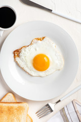Traditional breakfast. One fried egg on a white plate, toasts and coffee on a white table, top view, flat lay.