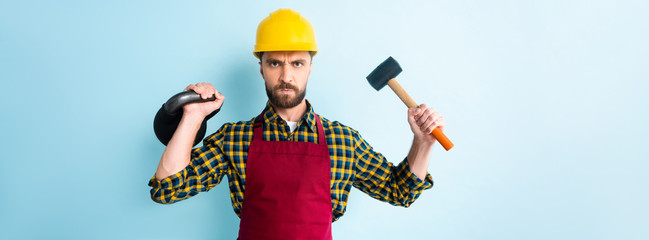 panoramic shot of angry workman holding hammer and dumbbell on blue