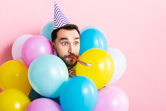 handsome man in party cap with party blower in mouth near balloons on pink