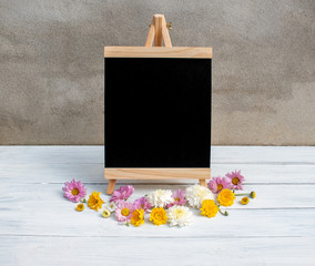 Blank chalkboard sign with wooden frame and wooden stand and many flowers in front of loft cement on white wooden background. Wedding Decoration. Flowers composition. Flat lay. Copy space.