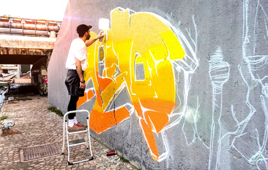 Street artist working on colored graffiti at public space wall - Modern art perform concept of urban guy painting live murales with yellow and orange aerosol color spray - Bright sunflare filter