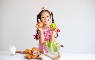 Lovely Asian little girl hold apples and smile with vegetables, milk and bread on table with white wall background.